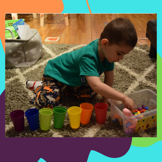 A 2-year-old child playing on the floor with colorful matching games, surrounded by various colored cups and small rubber fruits.