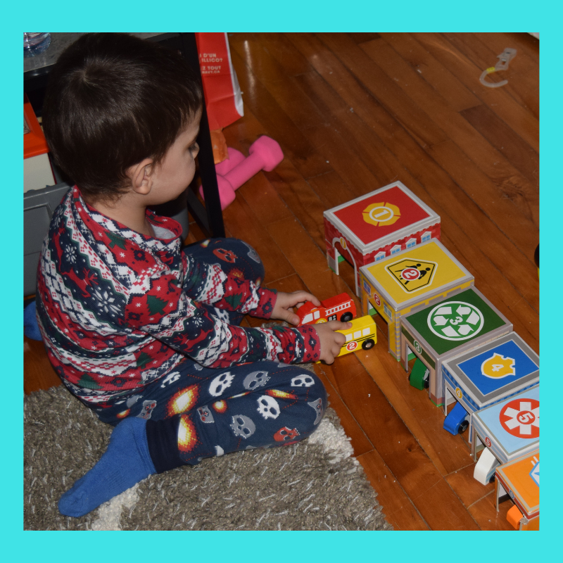 A 2-year-old child playing on the floor with numbered boxes of different sizes and numbered vehicles.